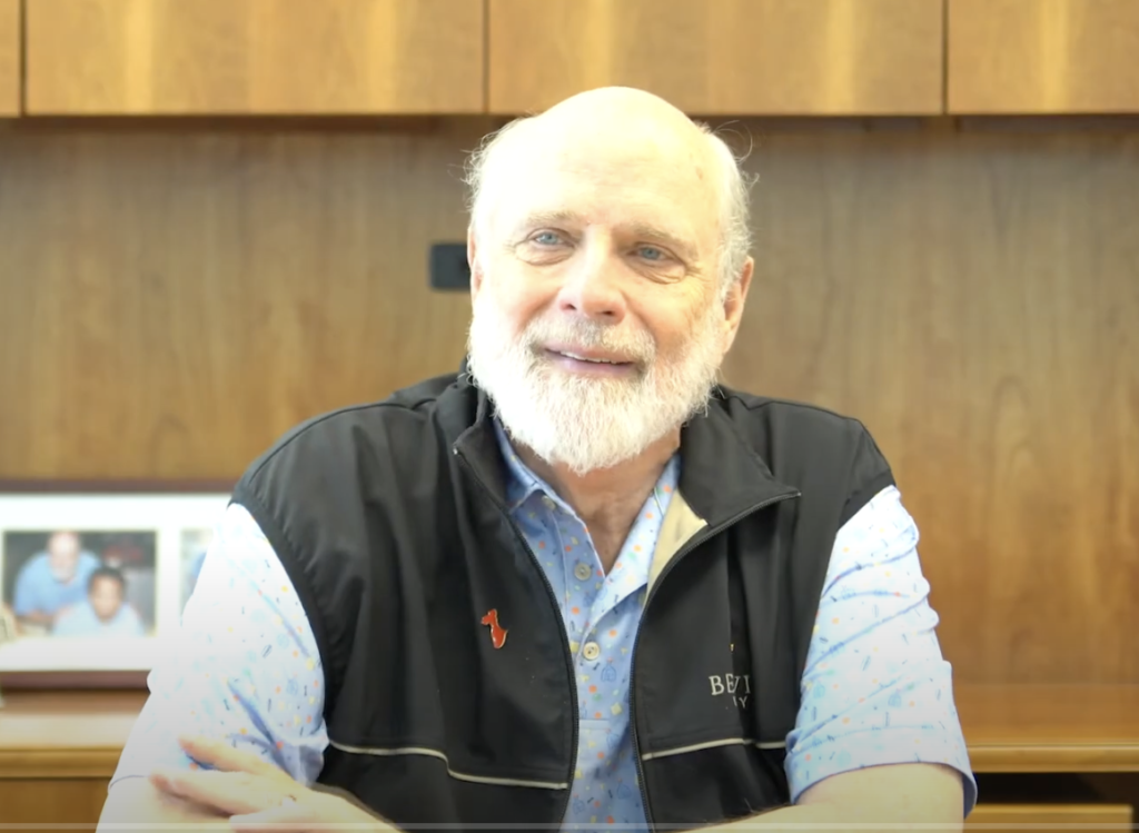 A man with a white beard is sitting at a desk wearing a black vest over a blue shirt. The desk has a wooden backdrop and a framed photo in the background.